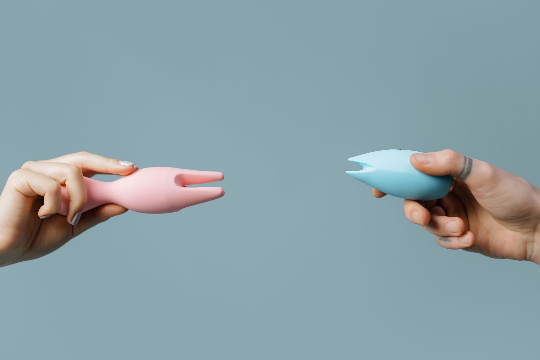 Zwei Hände halten rosa und blaue Silikonvibratoren vor einem Pastell-Hintergrund