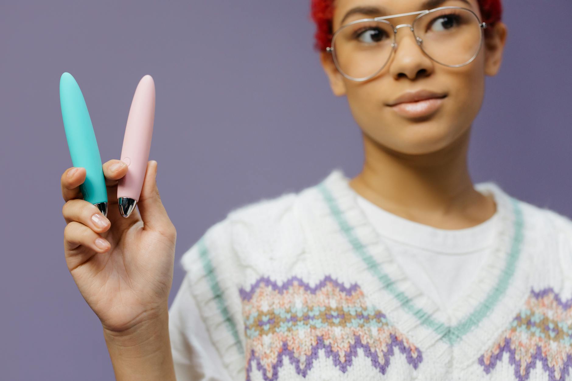 Eine junge Frau mit Brille hält zwei farbenfrohe Vibratoren gegen einen violetten Hintergrund.