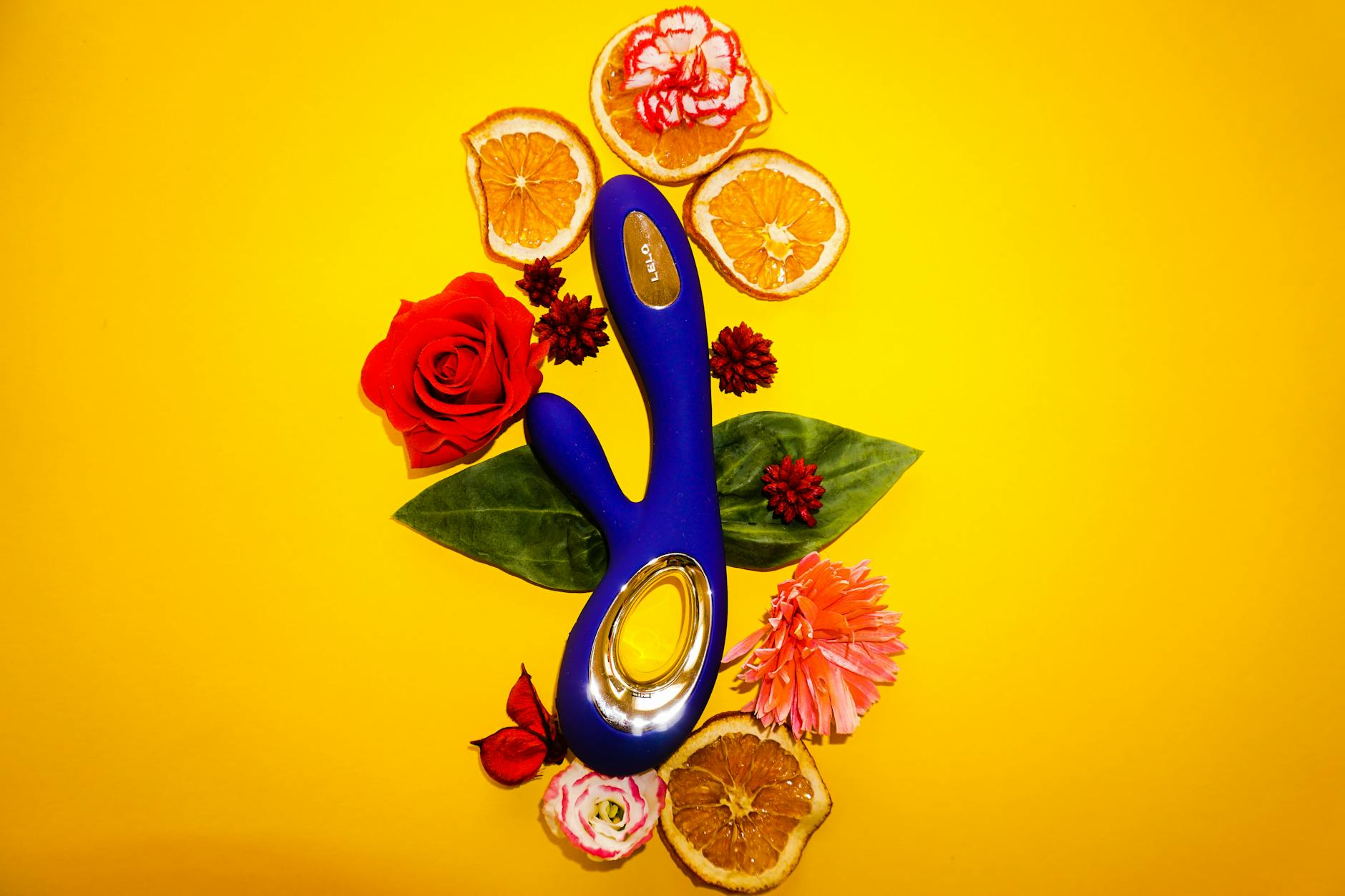 Ein blauer Lemon Vibrator neben Blumen und Orangenscheiben auf gelbem Hintergrund, das verschiedene Arten der Stimulation symbolisiert.